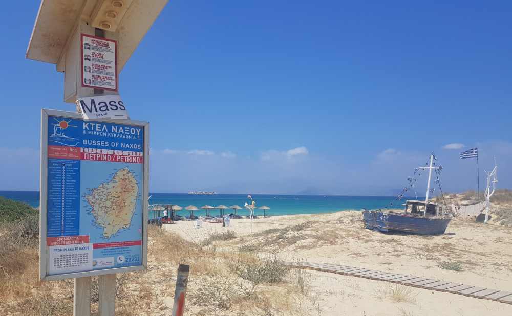 Naxos Greece: Travel Guide 2023 3 A photo of a bus timetable and map at a stop on a beach with the sea behind